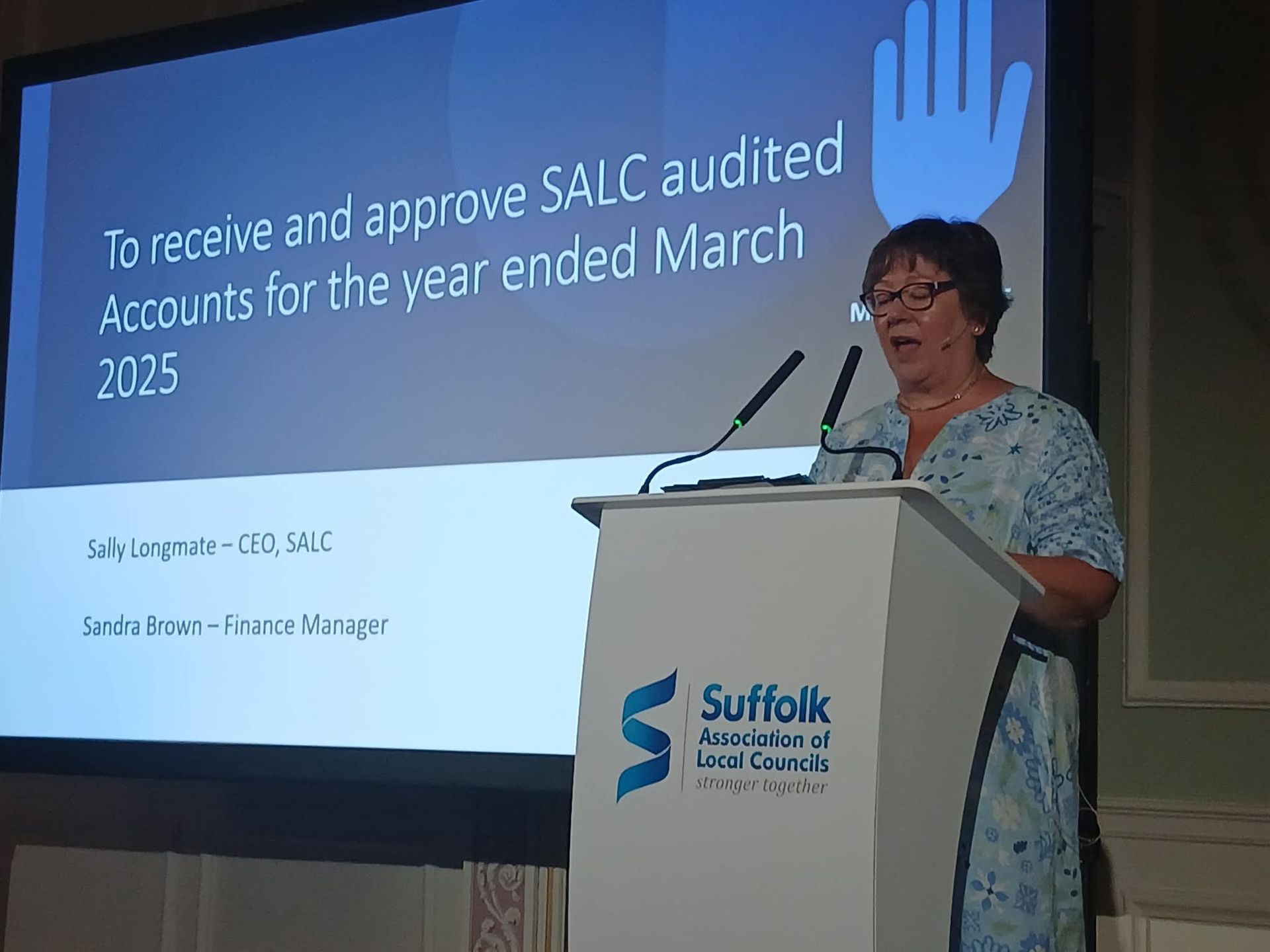 A woman speaks at a podium with a presentation screen behind her, discussing SALC audited accounts for 2025.