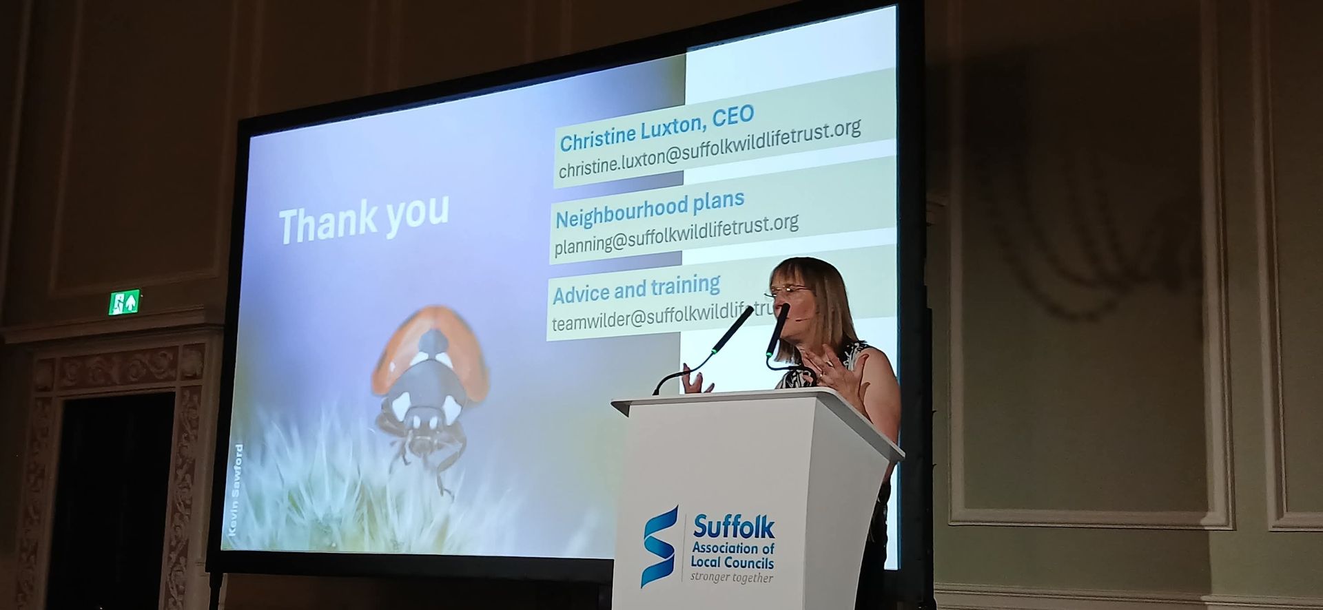 A speaker presents at a conference, with a slide showing a ladybug and contact information for a wildlife trust.