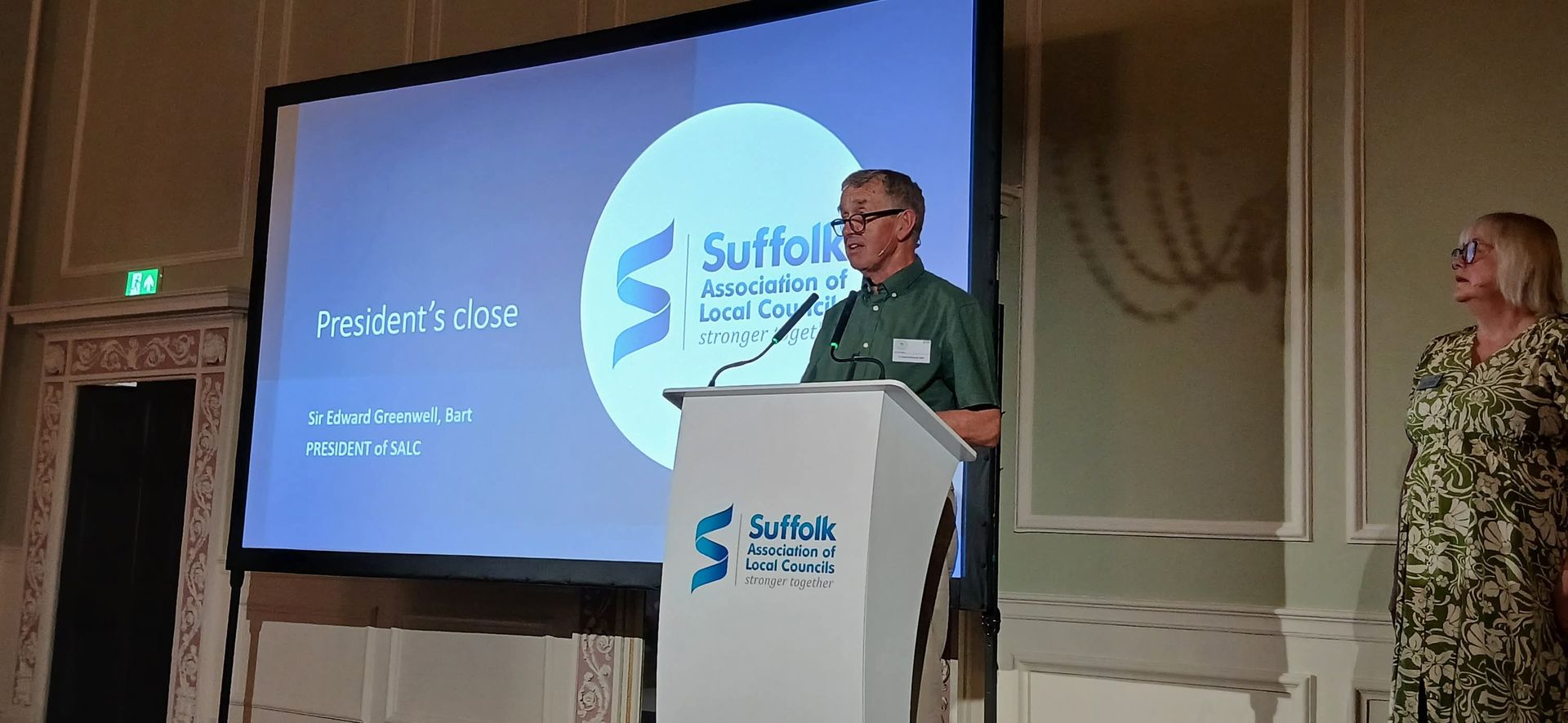 A speaker at a podium addresses an audience, with a presentation slide behind them displaying the Suffolk Association of Local Councils logo.