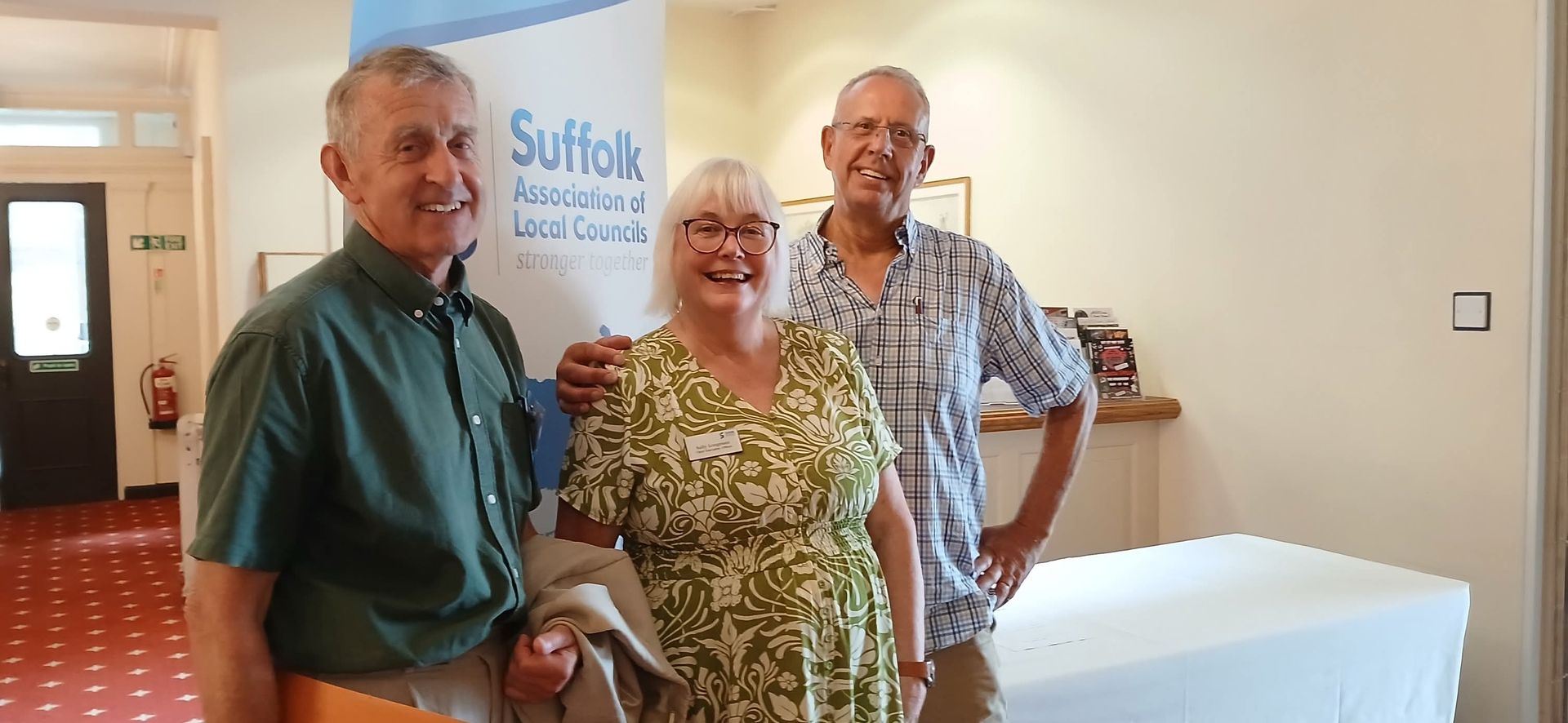 Three people pose together in a room with a banner for the Suffolk Association of Local Councils in the background.